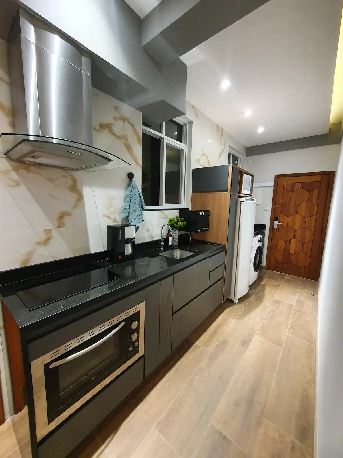 Cozinha compacta e alongada com bancada em granito preto, coifa, forno embutido e revestimento claro