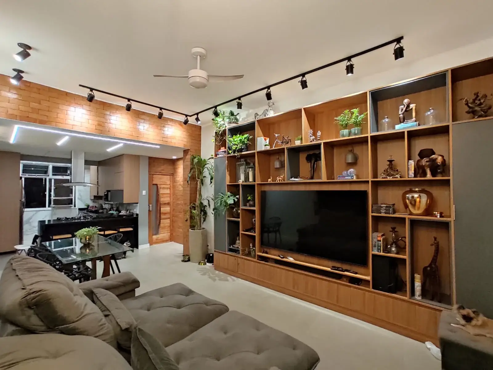 Sala de estar integrada à cozinha, com estante de madeira e televisão, iluminação em trilho, sofá cinza e ventilador de teto